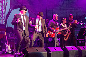 This image shows two singers, a guitarist, a sax player and a trumpet player at tyhe front of the stage in mid-performance as the Blues Brothers