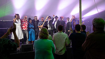 This image shows the full 11 piece band at the finale of a gig, centre stage. The are smiling and they have their arms around one another.