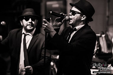 Two men dressed in black suits, fedoaa hats and sunglasses. They are singing on stage, as a Blues Brothers Tribute Act.